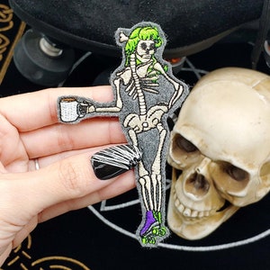 May include: An embroidered patch of a skeleton wearing roller skates and holding a cup of coffee. The skeleton has green hair and is wearing purple roller skates. The patch is on a black background.