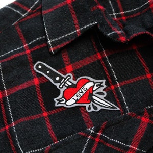 Puede incluir: Una camisa de franela a cuadros negros y rojos con un parche bordado de un corazón rojo y un cuchillo que dice "LOVE".