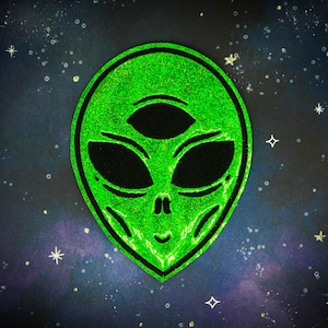 Pode incluir: Patch de rosto de alienígena verde com glitter, com três olhos pretos e contorno preto. O patch está sobre um fundo azul escuro com estrelas brancas.