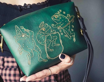 May include: Green crossbody bag with gold embroidered frog design. The bag features a rectangular shape with a zipper closure and a long, adjustable strap. The frog design is detailed with gold stitching and embellishments.