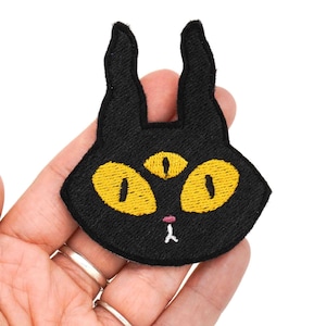 May include: Black embroidered iron-on patch of a cat with a third eye and yellow eyes.