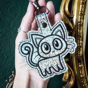 May include: A holographic silver cat keychain featuring black embroidered outlines of a cartoon cat with large eyes, pointed ears, and a curved tail. The keychain is attached to a metal lobster clasp by a matching holographic silver strap.