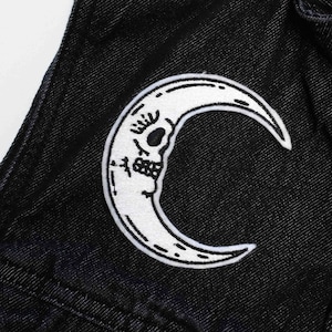 May include: A white crescent moon patch with a skull inside, sewn onto black denim fabric.