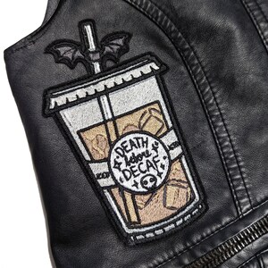 Puede incluir: Chaqueta de cuero negra con un parche de un vaso de café helado de dibujos animados con un murciélago en la parte superior y el texto "Death Before Decaf".