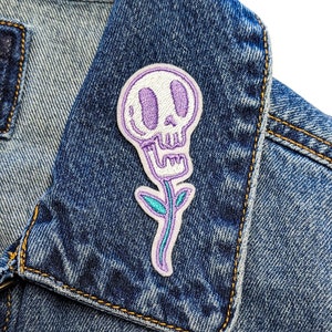 May include: A white skull with purple eyes and a purple stem and leaves on a denim jacket.