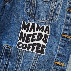 Op de afbeelding: Een spijkerjack met een witte patch waarop in zwarte letters "Mama Needs Coffee" staat.
