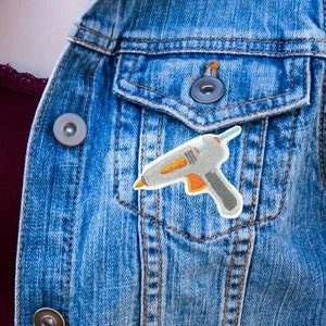 Puede incluir: Un chaleco vaquero con un parche de pistola de pegamento azul y naranja en el bolsillo delantero.