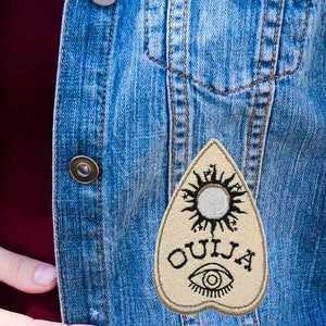 May include: A denim vest with a beige embroidered patch featuring a sun, eye, and the word "OUIJA" on a pointed shape.