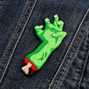 Op de afbeelding: Groen geborduurd zombiehand-patch met rood bloed dat druppelt van de afgehakte pols. De patch is genaaid op een blauw denim jack.