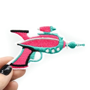 Pode incluir: Um remendo bordado de ferro de uma pistola de raios espaciais retro em rosa e turquesa.