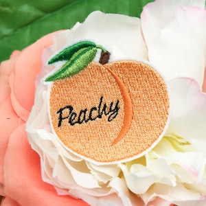 May include: Peach-shaped embroidered iron-on patch with the word "Peachy" in black thread. The patch is on a white and pink flower.