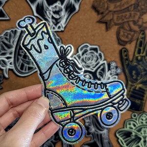 Puede incluir: Un parche de patín de ruedas de color plateado iridiscente con un detalle de hueso en la parte superior. El parche está hecho de una tela brillante y tiene un contorno negro.