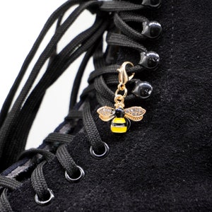 Puede incluir: Bota negra de gamuza con cordones negros y un dije de abeja dorada. La abeja tiene rayas amarillas y negras y brillantes de cristal transparente.