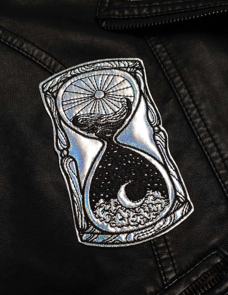 May include: A silver holographic iron-on patch featuring a detailed design of an hourglass with a sun and moon inside. The hourglass is surrounded by a decorative frame.