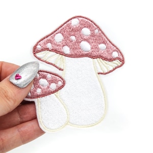Könnte beinhalten: Zwei gestickte Pilz-Patches in Rosa und Weiß. Der größere Pilz hat einen rosafarbenen Hut mit weißen Punkten und einen weißen Stiel. Der kleinere Pilz hat einen rosafarbenen Hut mit weißen Punkten und einen weißen Stiel.