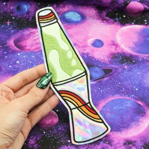 May include: A green and white lava lamp iron-on patch with a holographic rainbow design. The patch is on a purple and pink galaxy fabric background.
