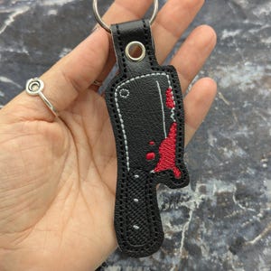 May include: Black leather keychain featuring an embroidered cleaver design with red blood splatter details. The keychain has a silver key ring and a stitched border. A unique accessory.