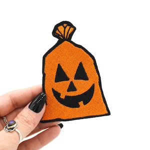 Op de afbeelding: Geborduurde oranje patch in de vorm van een Halloween-snoepzak met een pompoengezicht. De patch heeft een zwarte omtrek en details. De tas wordt tegen een witte achtergrond gehouden.