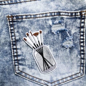 May include: Close-up of a light blue denim jean pocket with a decorative patch. The patch features a silver jar with black brush handles and brown brush heads. The jeans have distressed details.