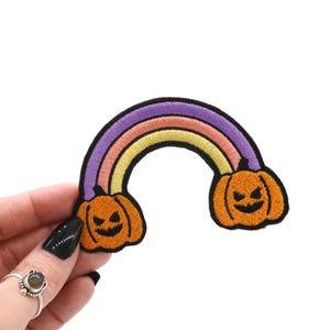 May include: Embroidered patch featuring a rainbow design with purple, pink, and yellow arches. Two orange jack-o'-lanterns with black facial features are at each end. The patch has a black border.