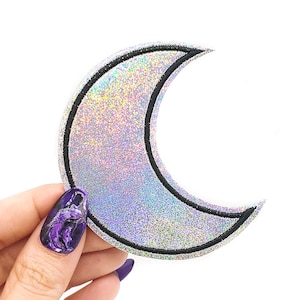 May include: A silver holographic crescent moon patch with a black outline. The patch is being held by a hand with purple nail polish.