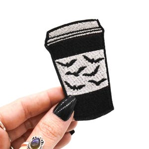 May include: Black embroidered patch in the shape of a coffee cup. The cup features a white rectangle with embroidered bat silhouettes. The top of the cup is white. The patch is held by a hand with black nail polish.