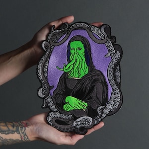 Op de afbeelding: Een geborduurde patch in zwart en grijs van de Mona Lisa met een octopuskop en tentakels. De patch is omgeven door een frame van zwarte en grijze octopus-tentakels.