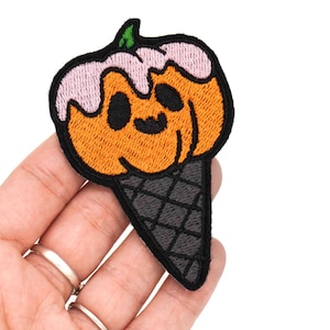Pode incluir: Um remendo bordado para passar a ferro com a forma de um cone de gelado de abóbora. A abóbora é laranja com um redemoinho rosa no topo e uma cara sorridente preta. O cone é preto e branco.