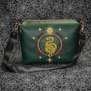 May include: Green crossbody bag with a gold zipper and black strap. The bag features an embroidered design of a snake inside a circle with a sun and stars. The background is a black and white spiderweb pattern.