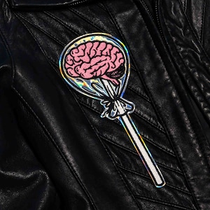 May include: A holographic iron-on patch of a pink brain on a lollipop stick. The patch has a rainbow iridescent finish.