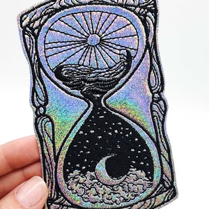 May include: A silver holographic iron-on patch with a black embroidered design of an hourglass with a sun and moon inside. The hourglass is surrounded by a decorative frame.