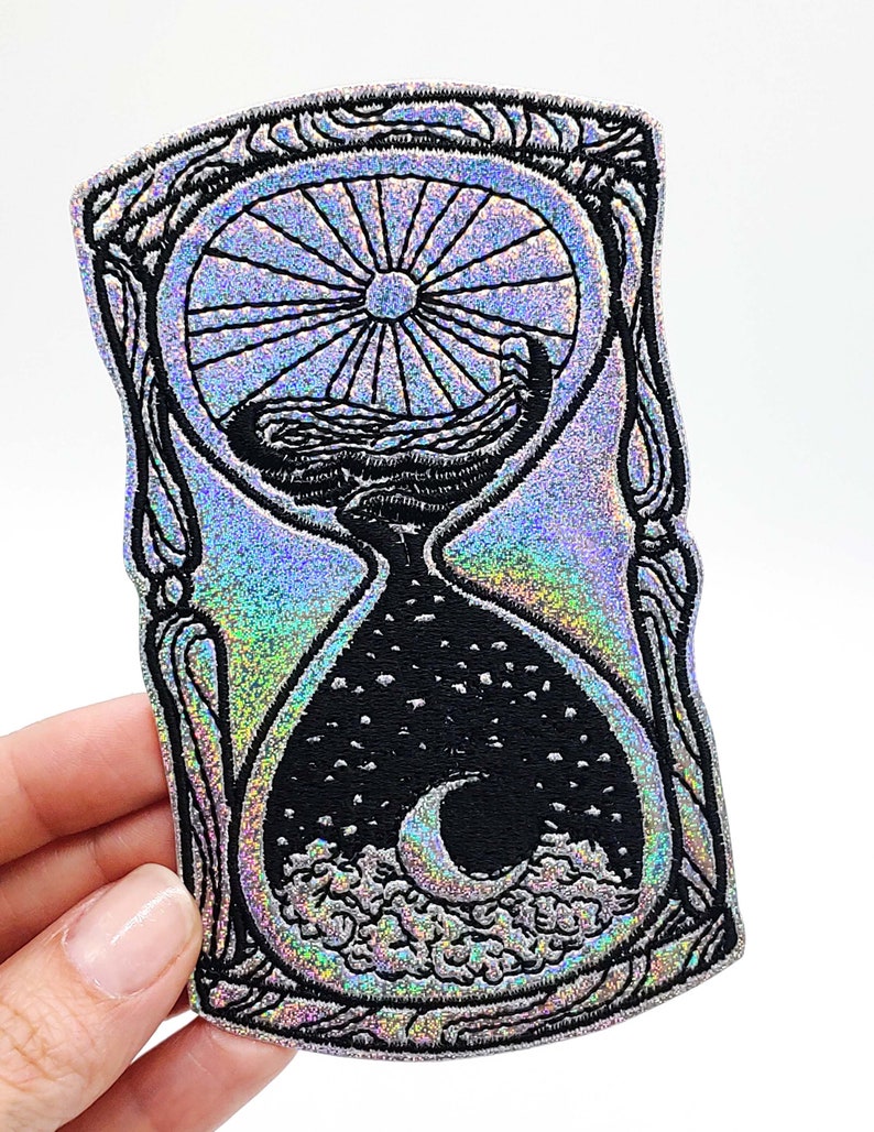 May include: A silver holographic iron-on patch with a black embroidered design of an hourglass with a sun and moon inside. The hourglass is surrounded by a decorative frame.
