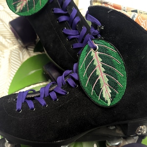 Può includere: Patini a rotelle neri con lacci viola e toppe a forma di foglia verde. I patini hanno ruote nere con lettere bianche.