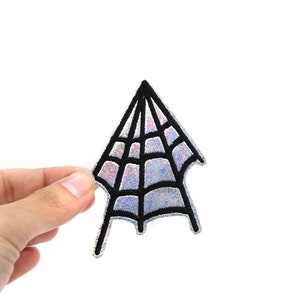 Może przedstawiać: Naszywka w kształcie pajęczyny w kolorze czarnym i srebrnym z efektem tęczowym. Naszywka ma kształt pajęczyny z ostrym wierzchołkiem i zaokrąglonym dołem. Pajęczyna jest obwiedziona czarną linią i wypełniona srebrnym materiałem z efektem tęczowym.