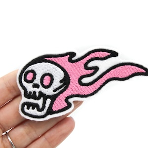 Peut inclure: Patch brodé rose et noir représentant un crâne de dessin animé avec des flammes. Le crâne a des yeux noirs et un contour noir. Les flammes sont roses avec un contour noir.