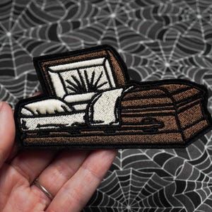 Könnte beinhalten: Gestickter Aufnäher mit einem braunen Sarg mit weißem Innenraum und offenem Deckel. Der Sarg hat einen schwarzen Rand und ist vor einem Spinnennetz-Hintergrund platziert. Ein Accessoire zum Thema Halloween oder Gothic.