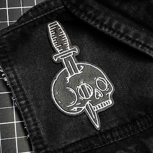 Könnte beinhalten: Ein schwarz-weißer Aufnäher mit einem Schädel, der von einem Dolch durchbohrt wird. Der Aufnäher ist auf eine schwarze Jeansjacke genäht. Der Schädel hat detaillierte Augenhöhlen und eine genähte Kontur.
