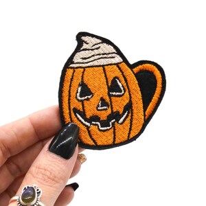Op de afbeelding: Geborduurde patch in de vorm van een pompoenmok met een jack-o'-lantern gezicht. De patch is oranje met zwarte en witte accenten. De mok heeft een zwart oor en een witte slagroom topping.