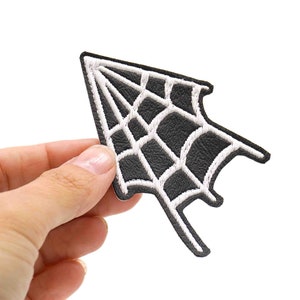 May include: Black felt spiderweb patch with white embroidered details. The patch is shaped like a spiderweb and has a pointed top.