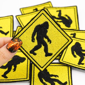 May include: A set of yellow and black embroidered iron-on patches featuring a silhouette of Bigfoot walking. The patches are shaped like a yellow warning sign with a black border.