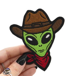 Könnte beinhalten: Gestickter Aufnäher mit einem grünen Alien, der einen braunen Cowboyhut und ein rotes Halstuch trägt. Der Alien hat große schwarze Augen und einen neutralen Gesichtsausdruck. Geeignet für Kleidung oder Accessoires.