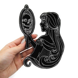 May include: Black embroidered iron-on patch featuring a woman looking into a mirror with a skull reflection. The woman has long flowing hair and is wearing a dress. The patch is outlined in white thread.