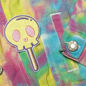 Pode incluir: Uma jaqueta de ganga tie-dye com um remendo de pirulito de caveira colorida e um botão de metal com o texto "Solid Blue" no lado esquerdo.