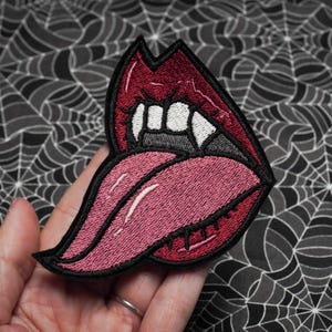 May include: Embroidered iron-on patch featuring a red mouth with fangs and a pink tongue sticking out.