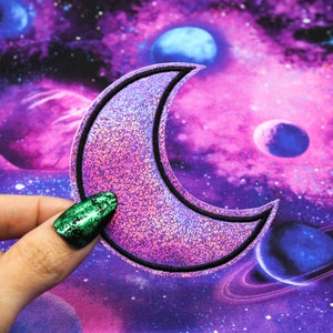 May include: A pink and purple glitter crescent moon patch with a black outline. The patch is on a purple and blue galaxy fabric background.