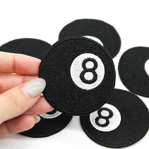 May include: Black embroidered patches featuring the number 8 in white, resembling a pool ball. The patches are round and made of a textured fabric. Several patches are visible, with one held by a hand.