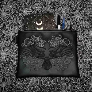 May include: Black zippered pouch with an embossed raven design, open to reveal a notebook with a crescent moon and stars, and pens. The pouch is on a black and white spiderweb patterned background.