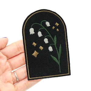 May include: A black embroidered patch with a white lily of the valley flower design and gold accents. The patch is shaped like an arch.