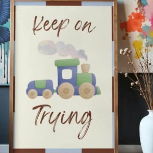 Peut inclure: Une affiche encadrée représentant un train jouet bleu et vert avec un panache de fumée. Le texte "Keep on Trying" est écrit en lettres cursives marron au-dessus et en dessous du train. Le cadre est en bois clair.