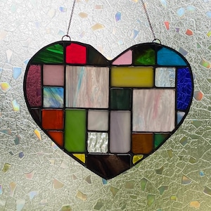 May include: A stained glass heart with a patchwork design of various colors hangs in front of a window. Several decorative glass bottles of different shapes and colors sit on the windowsill below the heart.